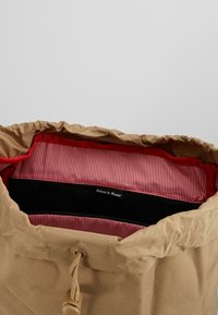 Beżowa torba na sznurek z otworem u góry, wyłożona czerwono-białą w paski podszewką oraz czarnym akcentem z etykietą "Witamy w Bristolu".