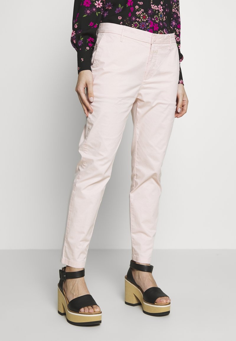 Femme portant un pantalon rose clair, un haut noir à fleurs et des sandales compensées noires à talons beiges épais, debout devant un fond uni.