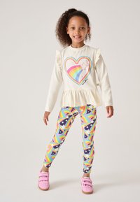 Crèmekleurige trui met ruches aan de mouwen, voorzien van een regenbooghartgrafiek. Kleurrijke legging met regenboogpatronen en sterren. Roze sneakers.