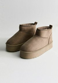 Bottines en daim marron à doublure en peluche, semelle à plateforme en caoutchouc plate et arrière légèrement surélevé. Détails de couture sur les côtés et petite languette de traction incluse.