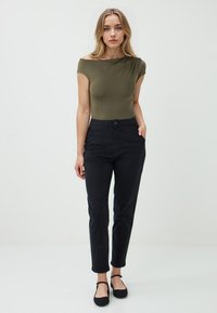 Olivgrüner, schulterfreier Body, kombiniert mit hoch taillierten schwarzen Hosen. Die Hosen verfügen über Taschen und einen Knopfverschluss. Schlichte schwarze Ballerinas.