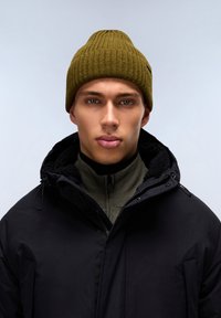Gorro de punto verde oliva con textura acanalada sobre un abrigo negro con cierre de cremallera y forro de forro polar, llevado sobre un suéter verde oliva con cremallera.