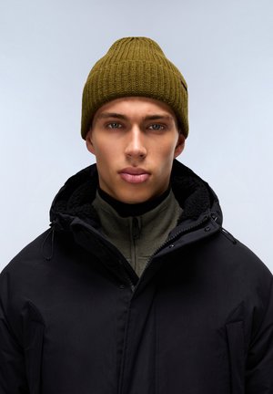 Cappello in maglia verde oliva con texture a coste, indossato sopra un cappotto nero con chiusura a zip e fodera in pile, sopra un maglione verde oliva con zip.