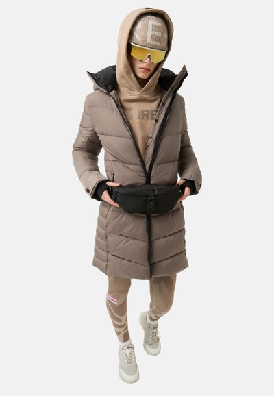 Steppmantel in Hellbraun mit schwarzen Akzenten, darüber getragen ein beiger Hoodie. Schwarze Gürteltasche an der Taille. Beigefarbene Leggings mit pinken Streifen und Muster.