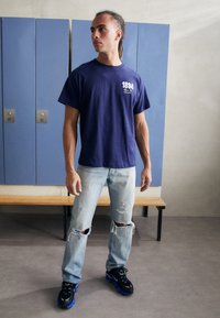 Navy crew-neck t-shirt with white graphic, light blue distressed jeans, and black sneakers with blue accents. Background features lockers.