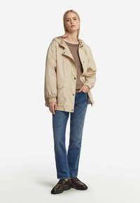 Beige, lätt jacka med huva, framfickor och elastiska muddarmar, bärs över en brun tröja och blå jeans; matchad med mörka skor.