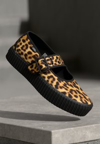 Leopárdmintás Vans slip-on cipő, fekete, vastag bordázott talppal és állítható csattal, egy kő felület fölött kiállítva.