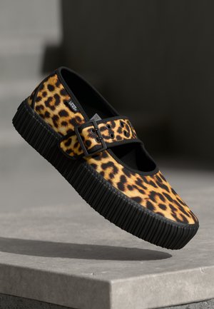 Zapatilla slip-on de Vans con estampado de leopardo, suela gruesa negra con estrías y correa ajustable con hebilla, expuesta sobre una superficie de piedra.