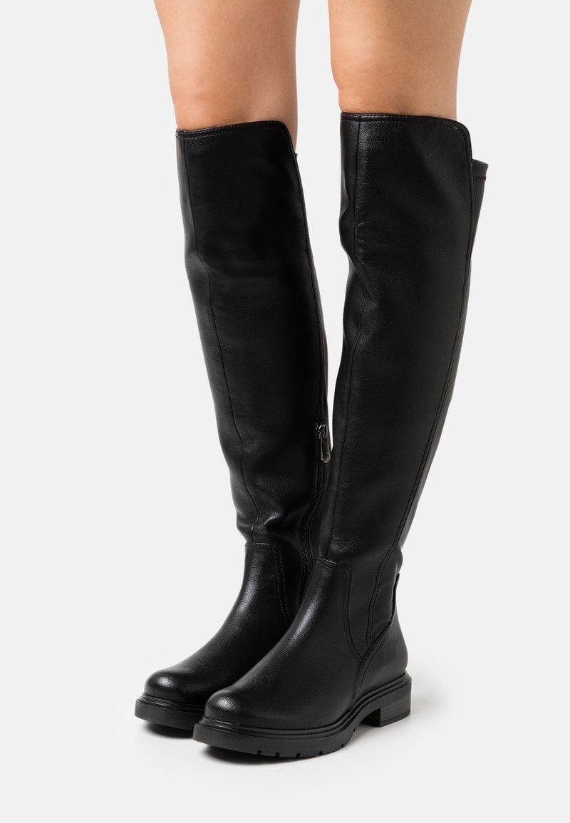 Bottes en cuir noires montant jusqu'aux genoux avec des fermetures éclair latérales, portées sur des jambes nues, avec des talons bas et épais et des bouts arrondis sur un fond neutre.