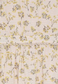 Blumengemusteter Stoff in sanftem Beige mit gelben und grauen Blumen. Strukturierter Gewebe mit kleinen Pompons entlang einer Naht.