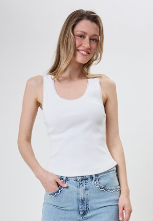 Ung kvinde smiler, iført en hvid ærmeløs ribbet top og lyseblå jeans med bølget kant på lommerne, stående foran en ensfarvet baggrund.