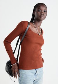 Jongeman met gevlochten haar, wearing een rustkleurige lange mouwen top, lichte spijkerbroek, zilveren ketting en armband, met een zwarte textuur schoudertas.