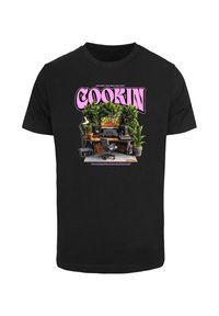 Sort bomulds T-shirt med et farverigt grafisk motiv med ordet "COOKIN" i pink, omgivet af et skrivebord med planter, højttalere og udstyr.