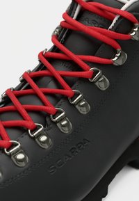 Schwarzer Scarpa Wanderschuh mit roten Schnürsenkeln, die durch silberne Metallösen gefädelt sind, auf einem weißen Hintergrund.