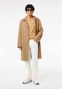 Cappotto trench marrone su una camicia beige, abbinato a pantaloni bianchi e sneakers bianche. Il modello indossa occhiali da sole e ha i capelli ricci.