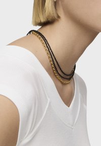 Tre collane a strati: una in oro, una con perline nere e una sottile catena nera, posate su un collo contro una camicia bianca.