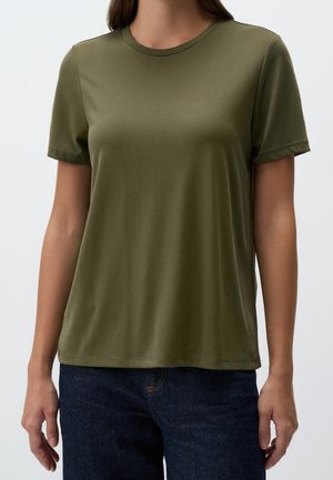 Vrouw draagt een olijfgroen T-shirt met korte mouwen en ronde hals en een donkerblauwe spijkerbroek, staand tegen een effen lichte achtergrond.