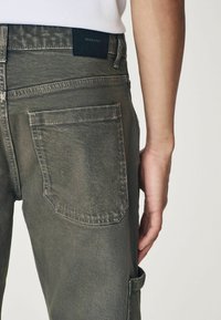 Jeanși din denim verde închis au un design ajustat, cusături vizibile și buzunare pe spate. Textura pare ușor uzată, cu un finisaj mat.