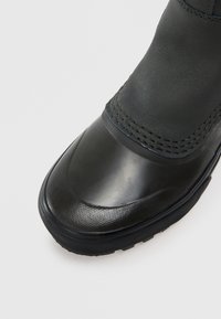 Primer plano de una bota negra impermeable que muestra la puntera texturizada, la costura, y la suela de goma gruesa sobre un fondo blanco.