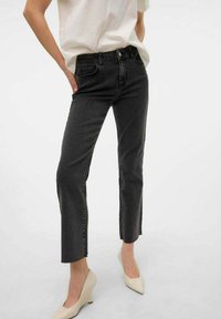 Jeans negros de pierna recta con cintura alta y dobladillo sin acabar. La tela parece tener una textura ligeramente rugosa. Combinados con zapatos claros y una blusa blanca suelta.