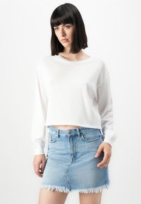 Kvinne med rett svart hår iført en hvit langermet cropped skjorte og et lyst blått slitt denim miniskjørt som står mot en hvit bakgrunn.