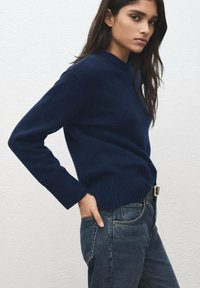 Temno modra pletena majica z okroglim izrezom in rebrastimi manšetami, kombinirana s temnimi denim kavbojkami. Majica je krojena po meri.