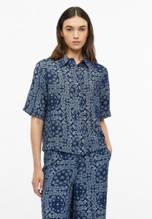 Femme portant une chemise à manches courtes bleue assortie et un pantalon avec des motifs cachemire et floraux blancs, debout avec une main dans la poche.