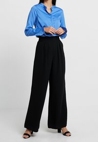 Woman wearing bright blue blouse with ruffled cuffs and wide-leg black trousers, standing with hands clasped at waist.