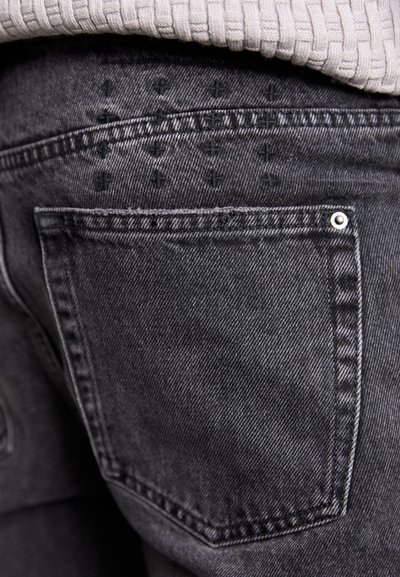 Des jeans en denim noir présentent une poche arrière avec des détails de couture et un clou en métal argenté. Le tissu a une texture lisse.