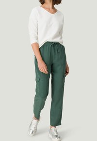 Mujer vestida con camisa blanca de manga larga, pantalones cargo verdes con cordón, y zapatillas plateadas metálicas, de pie sobre un fondo liso.