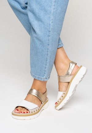 Feet wearing white and gold open-toe platform sandals with strap closures paired with cropped light blue jeans.