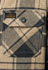 Sand- und dunkelblaue Karostoff mit strukturierter Oberfläche. Verfügt über eine Brusttasche mit einem Druckknopfverschluss und einem Etikett mit der Aufschrift "STANDARD ISSUE".