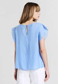 Blusa azzurra a maniche corte con finitura texturizzata, caratterizzata da un dettaglio a chiave sul retro e una forma morbida e fluida. Abbinata a pantaloni bianchi.