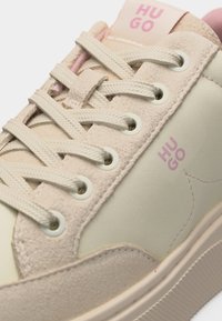 Κοντινό πλάνο μπεζ και ανοιχτό ροζ αθλητικού παπουτσιού με υφασμάτινα κορδόνια, πάνελ από σουέντ και δέρμα, και το λογότυπο "HUGO" στη γλώσσα και στην πλευρά.