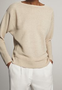 Maglione beige a coste con scollo a barca ampio, vestibilità rilassata e texture sottile. Abbinato a pantaloni bianchi, che mettono in risalto toni neutri morbidi.