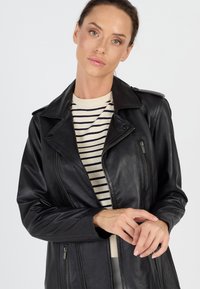 Schwarze Lederjacke mit Reißverschlusstaschen und Reverskragen, über einem gestreiften beigen Pullover getragen, der eine glatte Textur zeigt.