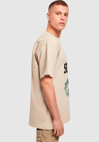 Beige bavlněné tričko s krátkým rukávem, na přední straně s výrazným černým textem a grafikou dolarové bankovky v zelené barvě. Rovný střih.