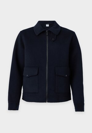 Jeune homme portant une veste noire, une chemise marron, un jean bleu clair et des chaussures beiges et noires, debout sur un fond blanc.