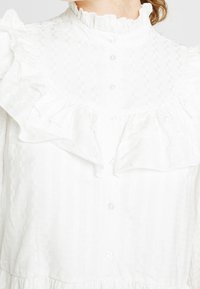 Vit blus med en texturerad mönster, volangkrage och knappstängning. Tyget är lättviktigt och har en subtil lyster.