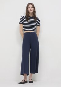 Pantaloni a gamba larga blu navy con bottone frontale, abbinati a un top corto a righe bianche e nere. Tessuto morbido e silhouette dritta. Ballerine nere.