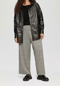Schwarze Kunstleder-Hemdbluse mit Knöpfen und zwei vorderen Taschen, kombiniert mit grauen karierter weite Hosen und schwarzen klobigen Slippern.