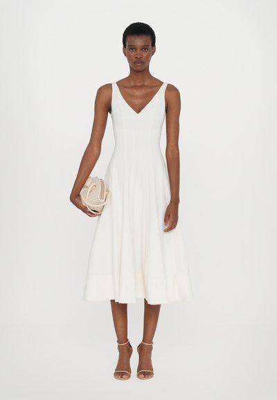 Witte midi-jurk met een V-halslijn, getailleerd lijfje en uitlopende zoom. Gladde stof, minimale ontwerpelementen, gecombineerd met een gestructureerde beige handtas.
