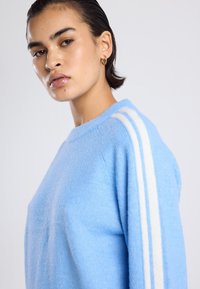 Pull léger bleu en matériau doux, présentant trois rayures verticales blanches sur l'épaule et un col rond.