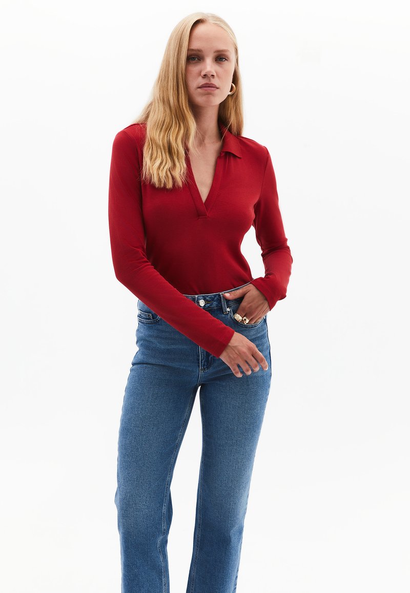 Rotes Langarmoberteil mit V-Ausschnitt und figurbetonter Passform, kombiniert mit blauen hoch taillierten Jeans. Glatte Stofftextur, minimale Hardwaredetails.