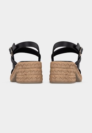 Par sorte sandaler med stropper, flettet tan plateauhæl og ankelspænder, vist bagfra på en ensfarvet baggrund.