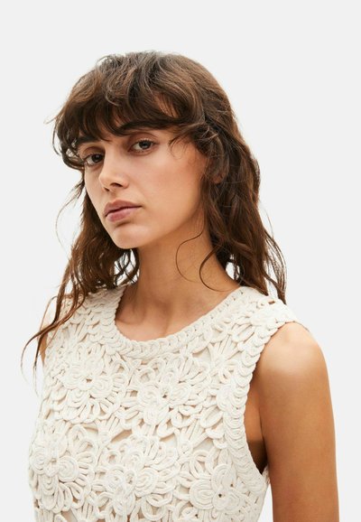 Joven con cabello castaño ondulado que lleva una blusa blanca sin mangas con un patrón texturizado de encaje floral, mirando ligeramente hacia un lado.