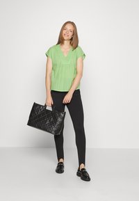 Top verde de mangas curtas com decote em V, combinado com leggings pretas. Acessório de uma mala de mão preta acolchoada. Textura matte e corte descontraído.