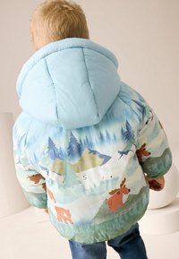 Veste de doudoune à capuche bleu clair avec un motif inspiré de la nature, mettant en scène des ours et des orignaux sur un paysage montagnard, texture matelassée.