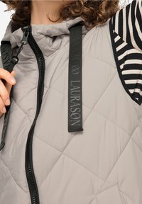 Jasnoszara pikowana kamizelka z czarnym zamkiem, z logo na pasku i w paskowaną czarno-białą koszulą pod spodem. Teksturowany, ocieplany materiał.