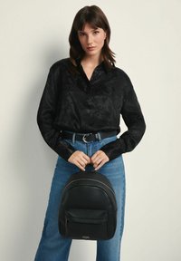 Czarny plecak z sztucznej skóry o zaokrąglonym kształcie, teksturowanej powierzchni i przedniej kieszeni. Modelka ma na sobie czarną koszulę w wzory i dżinsy z wysokim stanem.